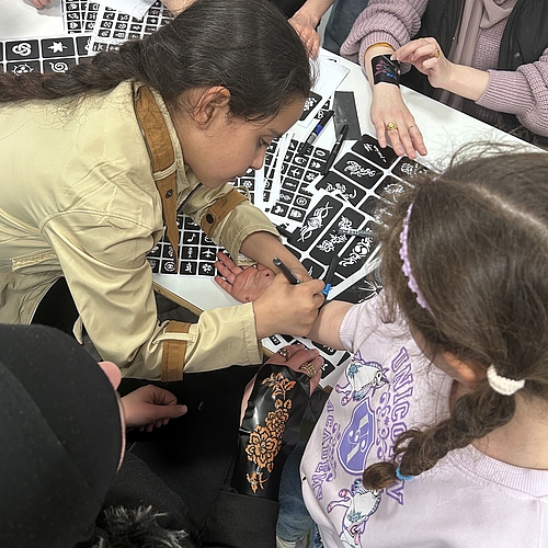 Mehrere Kinder stehen um einen Tisch mit zahlreichen Henna‑Tattoo‑Schablonen und malen sich gegenseitig Motive auf die Haut.