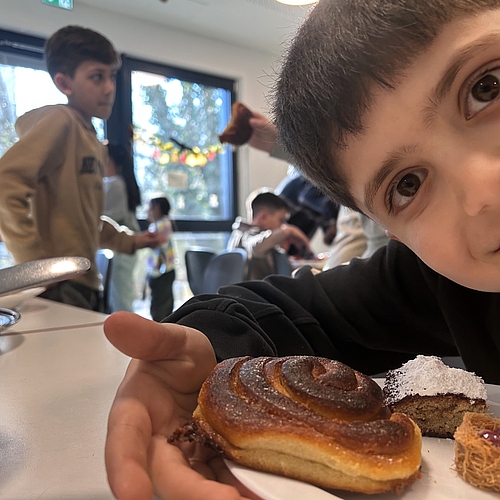 Ein Kind hält einen Teller mit süßem Gebäck, während im Hintergrund mehrere Kinder im Raum miteinander sprechen und essen.