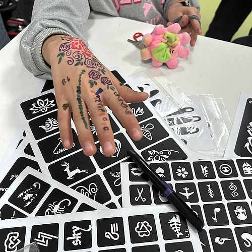 Ein Kind streckt die bunt bemalte Hand über einen Tisch voller Tattoo‑Schablonen und Stifte.